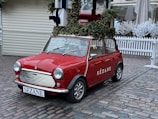 A vintage red Mini Cooper is parked on a cobblestone street. The car is decorated for the holidays with a wreath and garlands on top, along with large wrapped gift boxes. The brand name 'Sézane' is displayed on the side of the car, and a festive message is written on the windshield. Surrounding the scene are white picket fences and artificial snow-covered trees, adding to the winter ambiance.