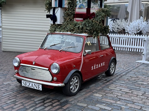 A vintage red Mini Cooper is parked on a cobblestone street. The car is decorated for the holidays with a wreath and garlands on top, along with large wrapped gift boxes. The brand name 'Sézane' is displayed on the side of the car, and a festive message is written on the windshield. Surrounding the scene are white picket fences and artificial snow-covered trees, adding to the winter ambiance.