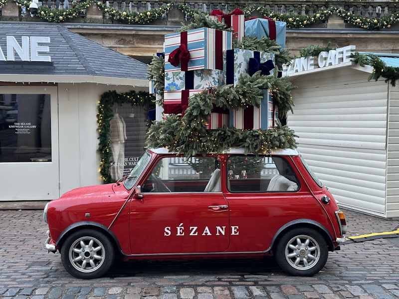 A red vintage Mini car is parked with a large stack of wrapped gift boxes and festive green garlands tied to its roof. The car is stationed on a cobblestone street in front of a caf&eacute; with holiday decorations, including a garland adorned with lights.