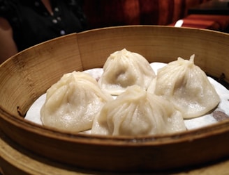 A bamboo steamer contains four well-crafted dumplings with delicate folds. These dumplings appear to be freshly steamed and sit on a layer of parchment paper. In the background, there is part of a person wearing a dark shirt visible.