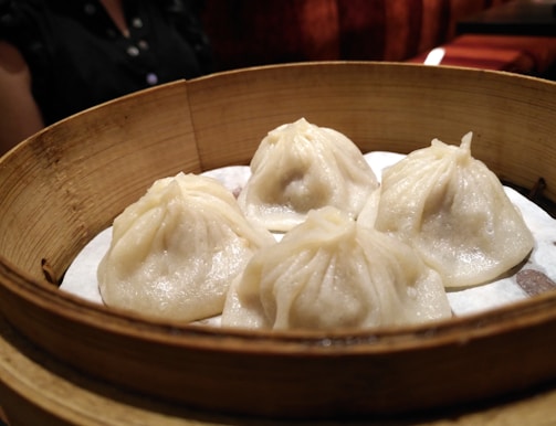 A bamboo steamer contains four well-crafted dumplings with delicate folds. These dumplings appear to be freshly steamed and sit on a layer of parchment paper. In the background, there is part of a person wearing a dark shirt visible.