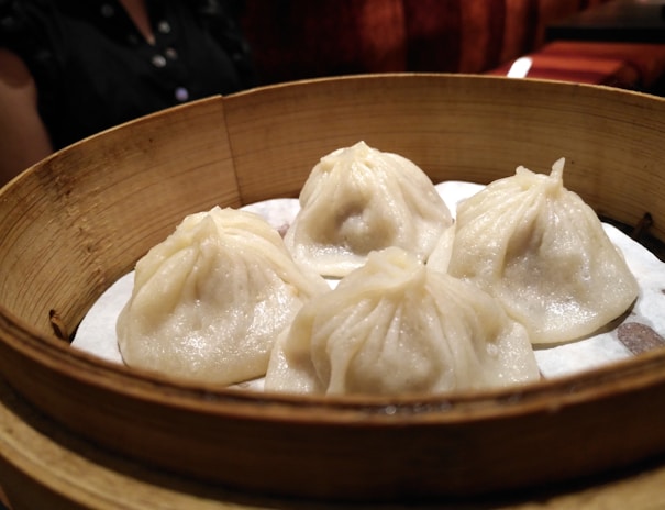 A bamboo steamer contains four well-crafted dumplings with delicate folds. These dumplings appear to be freshly steamed and sit on a layer of parchment paper. In the background, there is part of a person wearing a dark shirt visible.