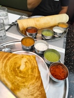A South Indian meal featuring two large plates with dosas and accompanying chutneys. The dosas are crisp and golden brown, served with an assortment of small bowls containing colorful chutneys and sambar. The setting includes a restaurant table with glass cups and cutlery.