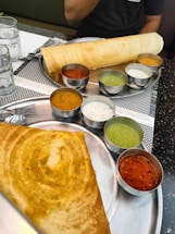 A South Indian meal featuring two large plates with dosas and accompanying chutneys. The dosas are crisp and golden brown, served with an assortment of small bowls containing colorful chutneys and sambar. The setting includes a restaurant table with glass cups and cutlery.