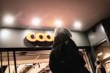 A person with gray hair is seen from behind walking into a building with a modern facade. Above the entrance, there is a brightly lit sign with the letters 'COO' clearly visible. The entrance is well-lit by overhead lighting, creating a warm and welcoming atmosphere.