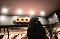 A person with gray hair is seen from behind walking into a building with a modern facade. Above the entrance, there is a brightly lit sign with the letters 'COO' clearly visible. The entrance is well-lit by overhead lighting, creating a warm and welcoming atmosphere.