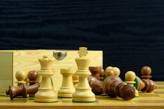 A collection of chess pieces, both white and brown, are arranged on a wooden chessboard. Some pieces are standing upright while others are toppled over. A wooden box with a metal clasp is in the background.