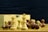 A collection of chess pieces, both white and brown, are arranged on a wooden chessboard. Some pieces are standing upright while others are toppled over. A wooden box with a metal clasp is in the background.