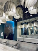 A modern bathroom with unique pendant light fixtures composed of overlapping wave-like structures hanging from the ceiling. The space has a mirrored wall reflecting these lights, creating an optical illusion. The area includes a long countertop with sleek stainless steel faucets and light-colored tiled walls.