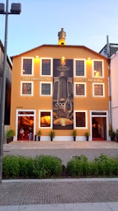 A vibrant orange building with white-trimmed windows and a sign reading 'Casa Portuguesa Pastel Bacalhau'. A large mural depicting culinary elements adorns the front, with small decorative plants positioned near the entrance. The building sits against a clear blue sky.