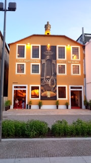 A vibrant orange building with white-trimmed windows and a sign reading 'Casa Portuguesa Pastel Bacalhau'. A large mural depicting culinary elements adorns the front, with small decorative plants positioned near the entrance. The building sits against a clear blue sky.
