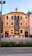 A vibrant orange building with white-trimmed windows and a sign reading 'Casa Portuguesa Pastel Bacalhau'. A large mural depicting culinary elements adorns the front, with small decorative plants positioned near the entrance. The building sits against a clear blue sky.