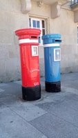 Two tall, cylindrical mailboxes are positioned side by side against a stone building with a window. One mailbox is bright red and the other is blue, both featuring a small slot at the top for mail deposit. They have signage with the word 'correio' and numbers on them, with a black base.