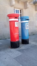 Two tall, cylindrical mailboxes are positioned side by side against a stone building with a window. One mailbox is bright red and the other is blue, both featuring a small slot at the top for mail deposit. They have signage with the word 'correio' and numbers on them, with a black base.