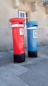 Two tall, cylindrical mailboxes are positioned side by side against a stone building with a window. One mailbox is bright red and the other is blue, both featuring a small slot at the top for mail deposit. They have signage with the word 'correio' and numbers on them, with a black base.