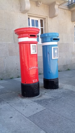 Two tall, cylindrical mailboxes are positioned side by side against a stone building with a window. One mailbox is bright red and the other is blue, both featuring a small slot at the top for mail deposit. They have signage with the word 'correio' and numbers on them, with a black base.