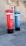 Two tall, cylindrical mailboxes are positioned side by side against a stone building with a window. One mailbox is bright red and the other is blue, both featuring a small slot at the top for mail deposit. They have signage with the word 'correio' and numbers on them, with a black base.
