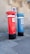 Two tall, cylindrical mailboxes are positioned side by side against a stone building with a window. One mailbox is bright red and the other is blue, both featuring a small slot at the top for mail deposit. They have signage with the word 'correio' and numbers on them, with a black base.