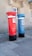 Two tall, cylindrical mailboxes are positioned side by side against a stone building with a window. One mailbox is bright red and the other is blue, both featuring a small slot at the top for mail deposit. They have signage with the word 'correio' and numbers on them, with a black base.