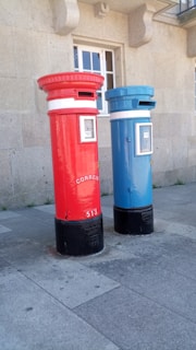 Two tall, cylindrical mailboxes are positioned side by side against a stone building with a window. One mailbox is bright red and the other is blue, both featuring a small slot at the top for mail deposit. They have signage with the word 'correio' and numbers on them, with a black base.