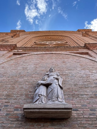 A stone sculpture features two figures, one adult and one child, standing against a large brick wall. The artwork is detailed, depicting flowing garments on both figures. Above them, architectural details of the building are visible with intricate design elements, under a bright blue sky with white clouds.