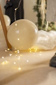A large white balloon rests on the floor amidst a string of small, glowing fairy lights. A piece of furniture is partially visible with a wooden leg supporting the balloon. In the background, a person in black attire, possibly holding a camera, stands near a decorated tree or plant with white fabric draped on the floor.