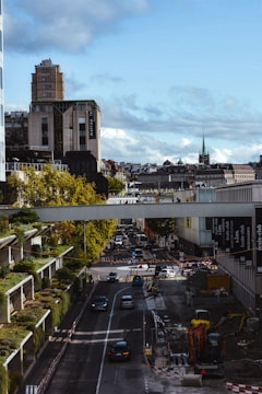 A cityscape featuring a busy urban street with ongoing construction work. Cars are driving along the road, and there is a construction vehicle and barriers present. Buildings surround the street, including a tall one on the left with a vertical garden and another with a distinctive spire in the background. The sky is partly cloudy with a blue tone.