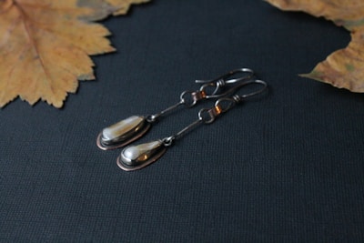 A pair of elegant, teardrop-shaped earrings made of silver with a polished finish. They are laid out on a dark textured surface, accompanied by dried autumn leaves, which add a natural, rustic feel to the composition.