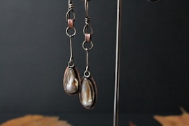 A pair of dangling earrings with iridescent white stones are hanging on a metal stand. The earrings have an elongated shape with a metallic finish, showcasing intricate looping details at the top. The background is a dark and plain surface, with some blurred elements at the bottom.
