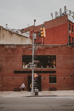 An urban street corner featuring a red brick building with large windows and a black door. There are multiple signs attached to a streetlight, including a stop sign and one-way signs. A woman is walking on the sidewalk adjacent to the building.