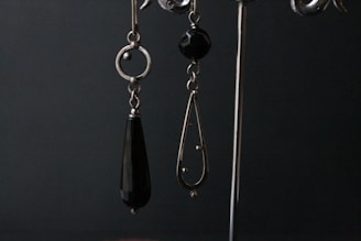 A pair of dangling earrings are displayed against a dark backdrop. The earring on the left is composed of a circular metal component with an elongated black teardrop-shaped bead hanging from it. The earring on the right features a black faceted bead at the top and a metal teardrop-shaped frame with small spherical accents and a larger spherical end piece.