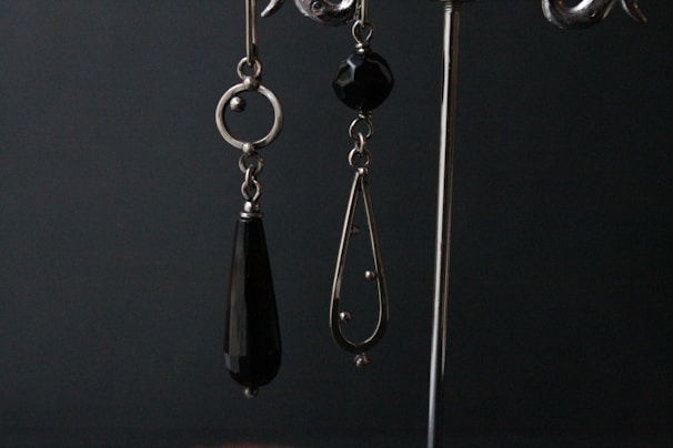 A pair of dangling earrings are displayed against a dark backdrop. The earring on the left is composed of a circular metal component with an elongated black teardrop-shaped bead hanging from it. The earring on the right features a black faceted bead at the top and a metal teardrop-shaped frame with small spherical accents and a larger spherical end piece.