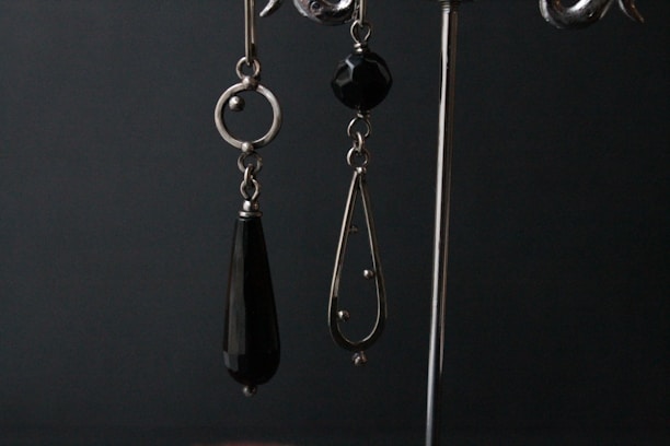 A pair of dangling earrings are displayed against a dark backdrop. The earring on the left is composed of a circular metal component with an elongated black teardrop-shaped bead hanging from it. The earring on the right features a black faceted bead at the top and a metal teardrop-shaped frame with small spherical accents and a larger spherical end piece.