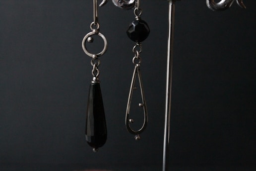 A pair of dangling earrings are displayed against a dark backdrop. The earring on the left is composed of a circular metal component with an elongated black teardrop-shaped bead hanging from it. The earring on the right features a black faceted bead at the top and a metal teardrop-shaped frame with small spherical accents and a larger spherical end piece.