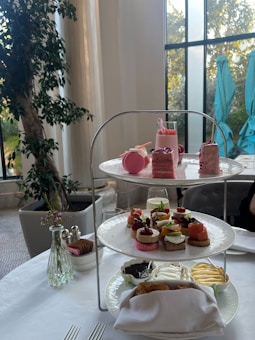 A three-tiered stand on a white table elegantly displays a selection of desserts and small sandwiches. On the top tier, there are pink macarons, pastries, and a small glass with a pink drink. The middle tier features a variety of finger sandwiches with toppings like salmon and caprese. The bottom tier includes small bowls of spreads and a napkin with some wrapped items. In the background, indoor plants and tall windows allow natural light to fill the room.