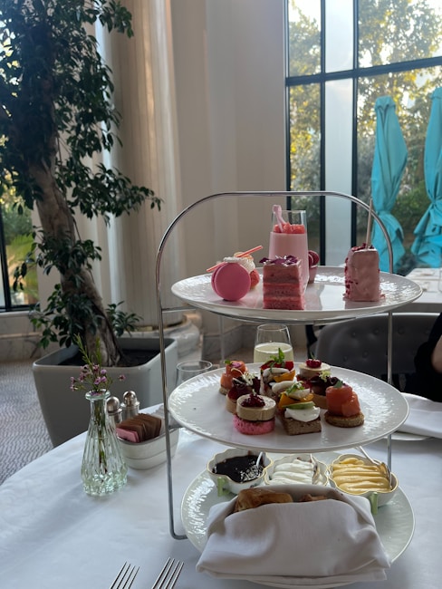 A three-tiered stand on a white table elegantly displays a selection of desserts and small sandwiches. On the top tier, there are pink macarons, pastries, and a small glass with a pink drink. The middle tier features a variety of finger sandwiches with toppings like salmon and caprese. The bottom tier includes small bowls of spreads and a napkin with some wrapped items. In the background, indoor plants and tall windows allow natural light to fill the room.