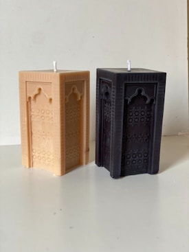 Two rectangular candles featuring intricate geometric patterns are placed side by side. The left candle is a light beige color, while the right one is a dark brown or black. The surface they rest on appears to be a simple, light-colored table, and the background is a plain wall.