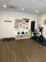 A fitness room featuring a variety of workout equipment neatly organized. The wall has a pegboard displaying exercise bands, foam rollers, and ropes. Below the board, there are kettlebells and dumbbells on shelves. An exercise bike is positioned to the right, with a step platform on the left.