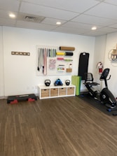 A fitness room featuring a variety of workout equipment neatly organized. The wall has a pegboard displaying exercise bands, foam rollers, and ropes. Below the board, there are kettlebells and dumbbells on shelves. An exercise bike is positioned to the right, with a step platform on the left.