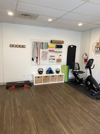 A fitness room featuring a variety of workout equipment neatly organized. The wall has a pegboard displaying exercise bands, foam rollers, and ropes. Below the board, there are kettlebells and dumbbells on shelves. An exercise bike is positioned to the right, with a step platform on the left.