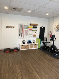 A fitness room featuring a variety of workout equipment neatly organized. The wall has a pegboard displaying exercise bands, foam rollers, and ropes. Below the board, there are kettlebells and dumbbells on shelves. An exercise bike is positioned to the right, with a step platform on the left.