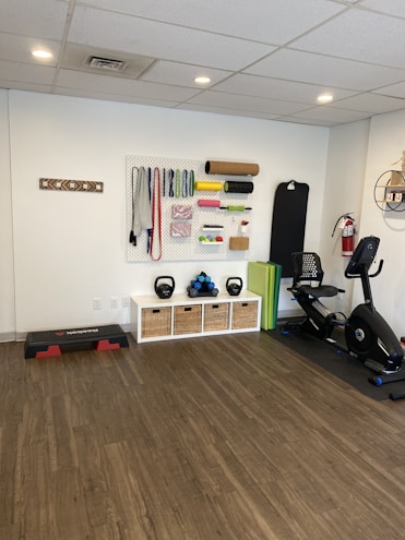 A fitness room featuring a variety of workout equipment neatly organized. The wall has a pegboard displaying exercise bands, foam rollers, and ropes. Below the board, there are kettlebells and dumbbells on shelves. An exercise bike is positioned to the right, with a step platform on the left.