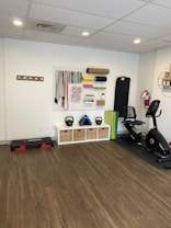 A fitness room featuring a variety of workout equipment neatly organized. The wall has a pegboard displaying exercise bands, foam rollers, and ropes. Below the board, there are kettlebells and dumbbells on shelves. An exercise bike is positioned to the right, with a step platform on the left.