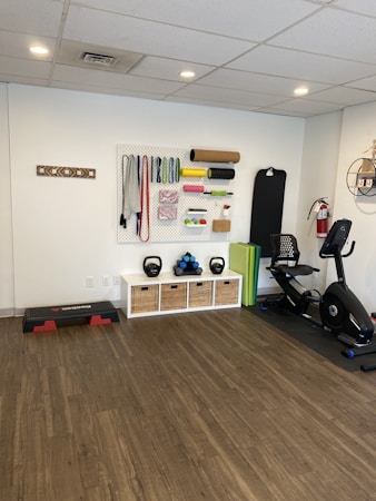 A fitness room featuring a variety of workout equipment neatly organized. The wall has a pegboard displaying exercise bands, foam rollers, and ropes. Below the board, there are kettlebells and dumbbells on shelves. An exercise bike is positioned to the right, with a step platform on the left.