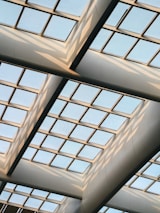A modern architectural ceiling design with a grid of large square glass panels, allowing natural light to filter through. The structure features intersecting metal beams and creates a pattern of shadows and reflections across the glass.