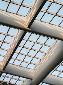 A modern architectural ceiling design with a grid of large square glass panels, allowing natural light to filter through. The structure features intersecting metal beams and creates a pattern of shadows and reflections across the glass.