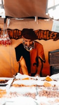 A vendor wearing a beret and traditional clothing is arranging a variety of nuts and dried fruits displayed in bags on a market stall. The background has a wooden sign with handwritten words. Sunlight streams through an opening, casting shadows.