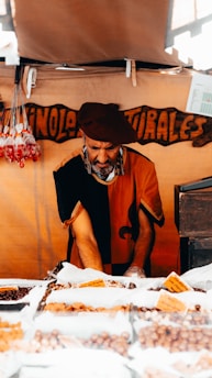 A vendor wearing a beret and traditional clothing is arranging a variety of nuts and dried fruits displayed in bags on a market stall. The background has a wooden sign with handwritten words. Sunlight streams through an opening, casting shadows.