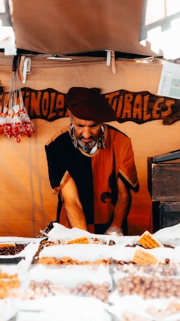 A vendor wearing a beret and traditional clothing is arranging a variety of nuts and dried fruits displayed in bags on a market stall. The background has a wooden sign with handwritten words. Sunlight streams through an opening, casting shadows.
