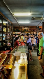 A small shop or market interior bustling with activity, featuring several people interacting at the counter. The shelves are stocked with a variety of goods, including bottles and packaged items. Overhead lighting brightens the space, and a ceiling fan can be seen.